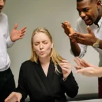 Seorang wanita tampak tertekan di kantor sementara rekan-rekannya berargumen di belakangnya, menggambarkan konflik di tempat kerja